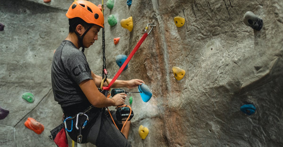 Accidentally dropped boulder in wrong hole in the distortion world - From below of side view of Asian male specialist in helmet drilling hole in blue climbing hold while hanging on safety rope near bouldering wall