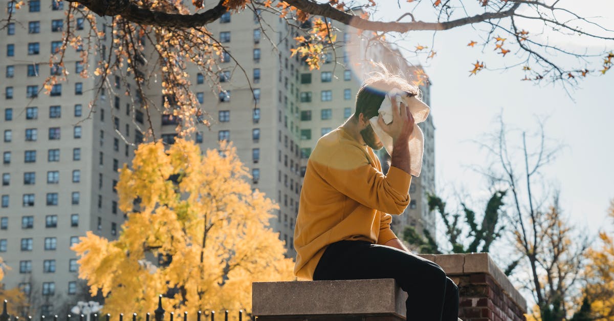 Autumn season challenge - Side view unrecognizable male in orange hoodie sitting on brick border in city park and wiping sweat from forehead with towel