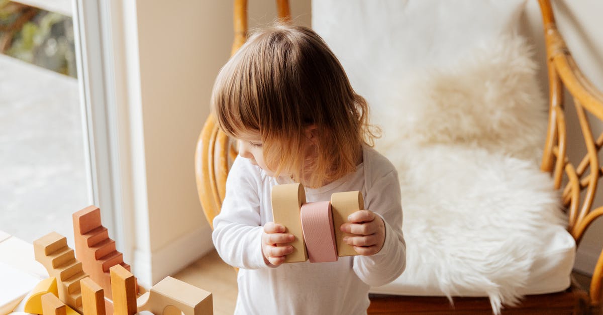 Block rotation preview box - From above of cute little child in white wear holding wood blocks for building while standing near wicker armchair