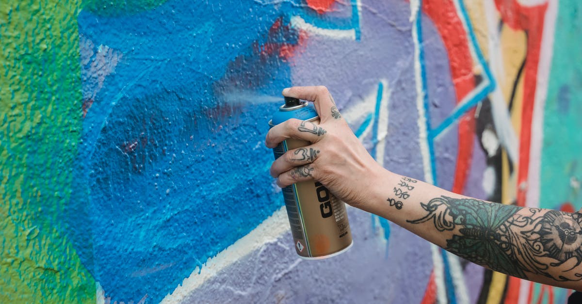 Can't Create Riot Account - Crop unrecognizable tattooed painter spraying blue paint from can on multicolored wall with creative graffiti while standing on street in city