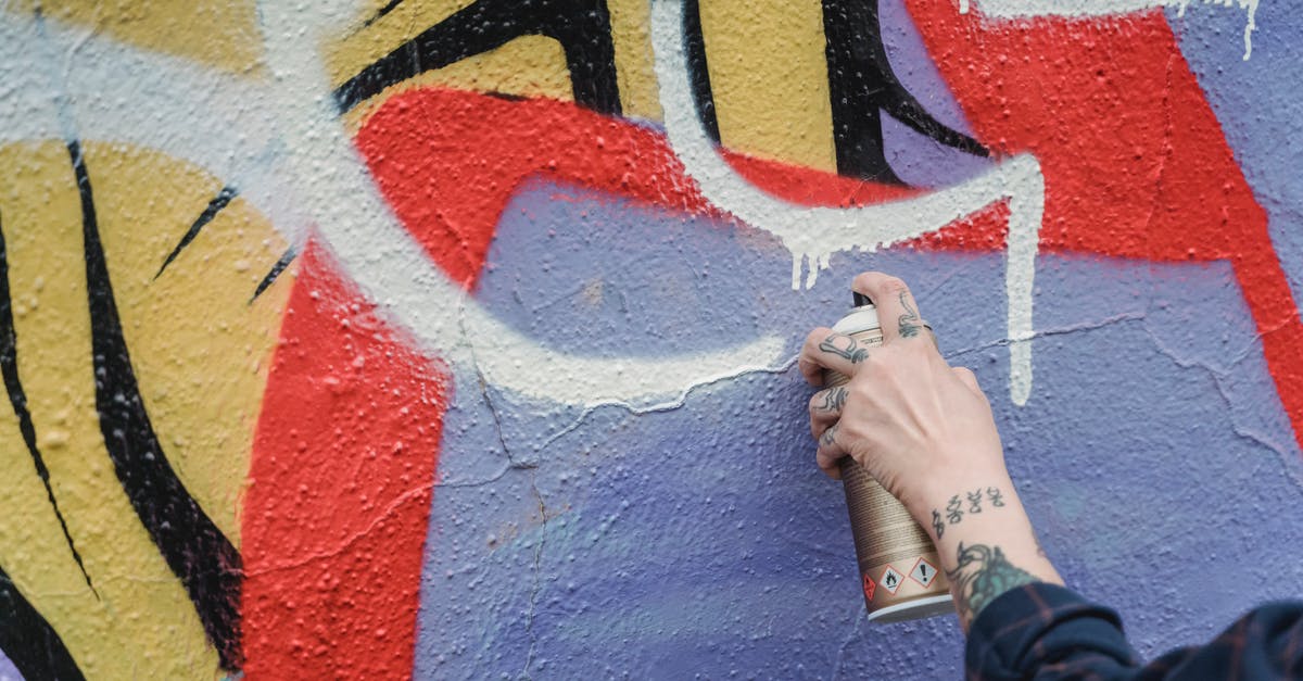 Can't Create Riot Account - Hand of crop anonymous tattooed person spraying white paint from can on colorful wall while standing on street of city