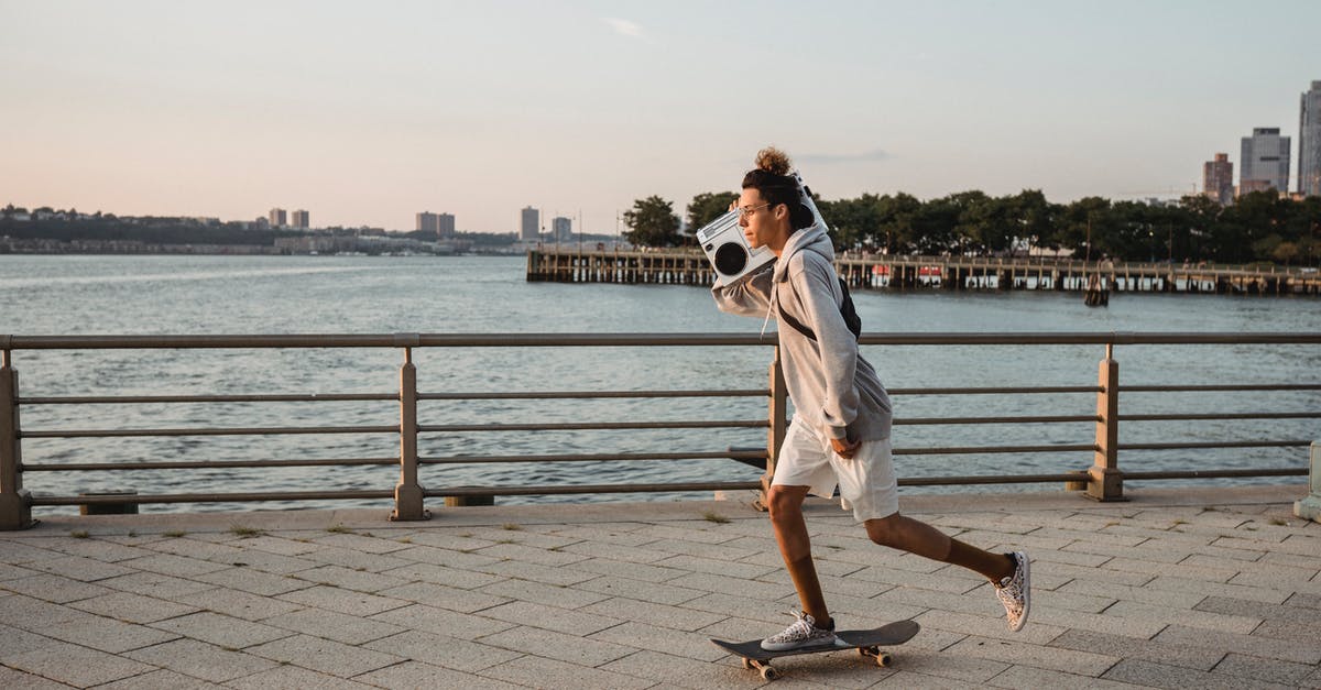 Can't hear any sound on True Skate - Side view full length male in casual clothes skating on city promenade and carrying retro tape recorder on shoulder in evening light