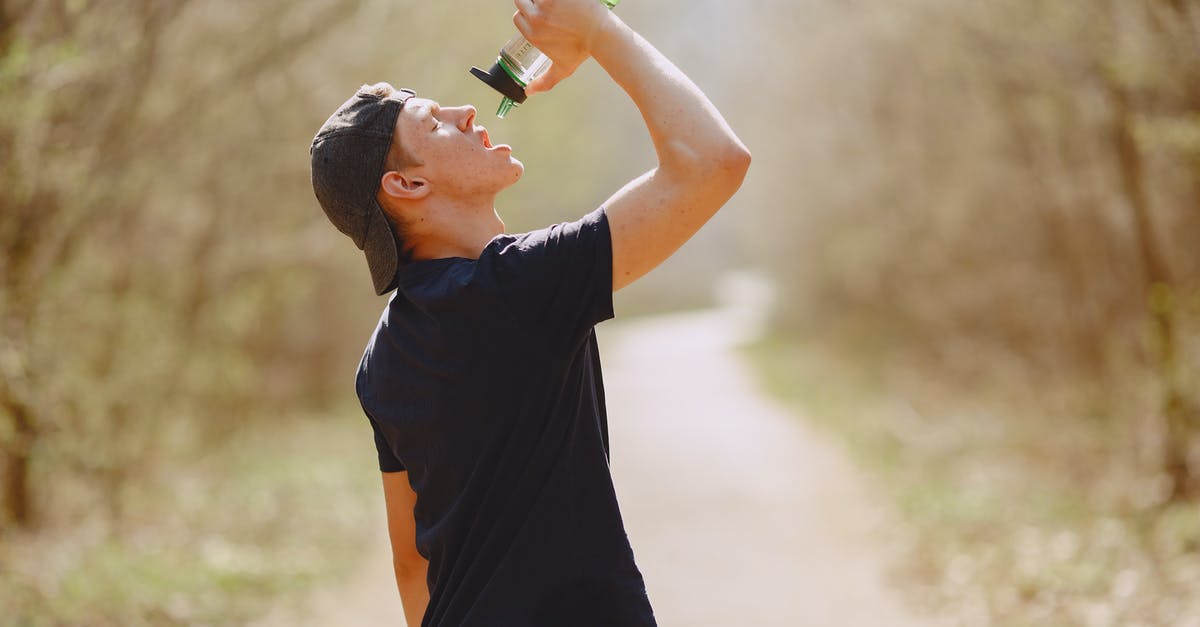 Can't run any program in - Side view of young male athlete wearing sports clothes and cap drinking water from plastic bottle on blurred background of forest during running and workout