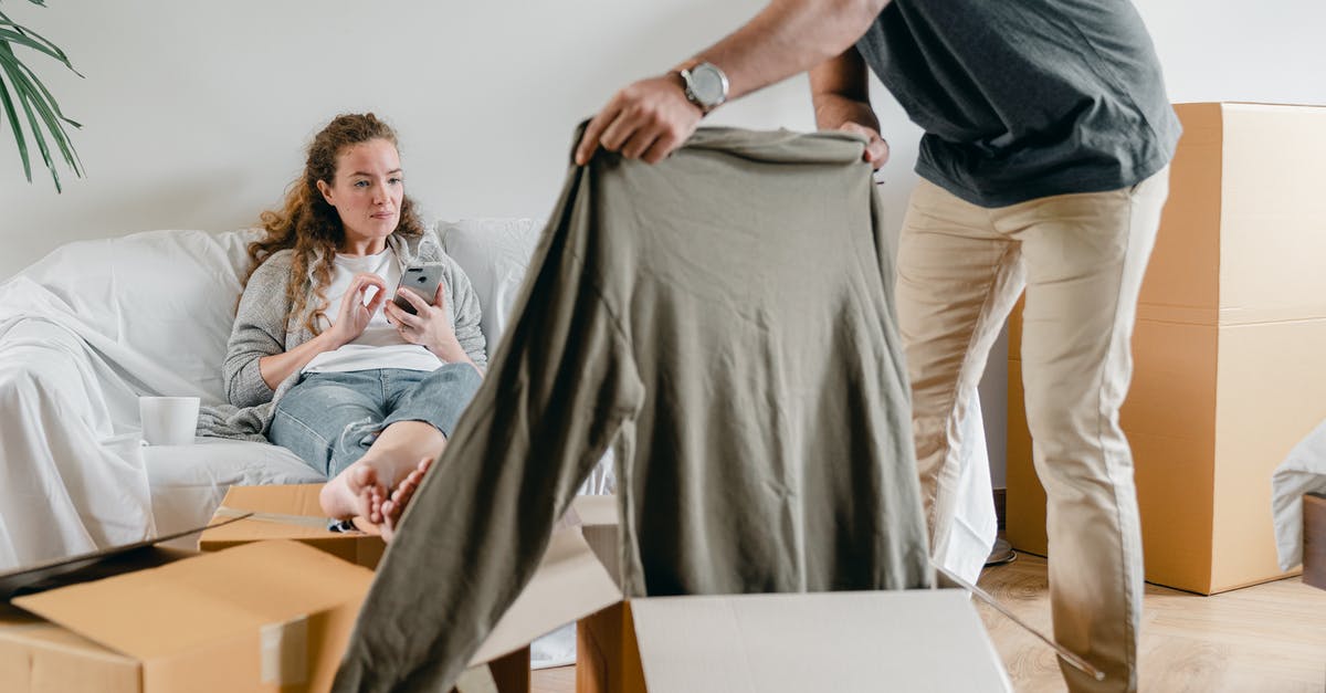 Can I get a new cracker? - Crop anonymous male in watch getting polo neck out of cardboard box while standing near pensive barefoot girlfriend sitting with crossed legs on comfortable couch after moving into new flat