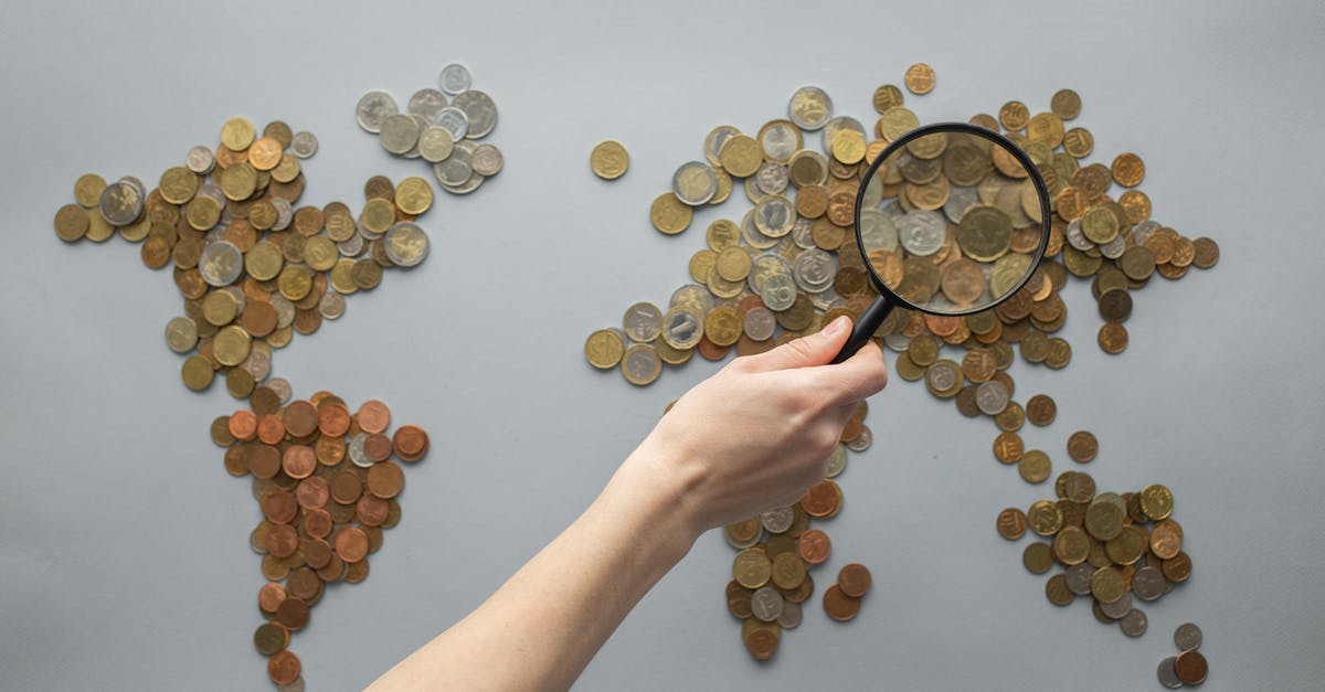 Can you make money from your settlements? - Top view of crop unrecognizable traveler with magnifying glass standing over world map made of various coins on gray background