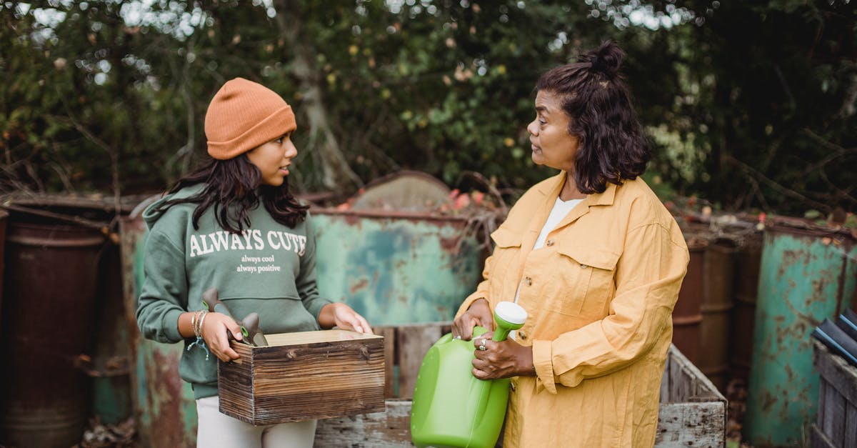 Can’t talk to my teammates - Ethnic woman with watering can speaking with teen near metal containers while looking at each other on farm