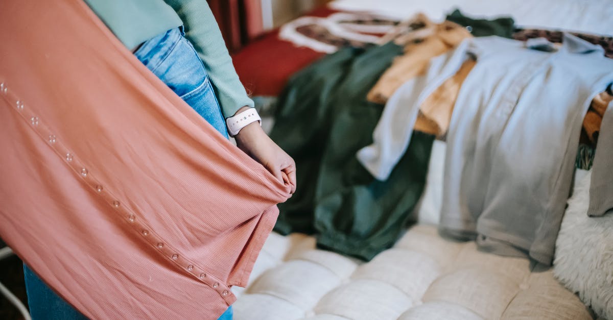 Do I have to select new quirks? - Crop anonymous woman in casual clothes demonstrating pink dress while selecting clothes while standing in bright bedroom near couch and bed with wardrobe at home