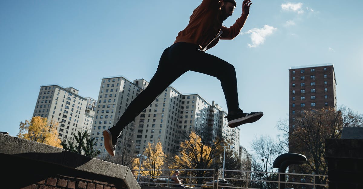 Do jump stunts benefit you? - Side view energetic young male tracer in casual clothes jumping from brick fence against modern city buildings on clear sunny day