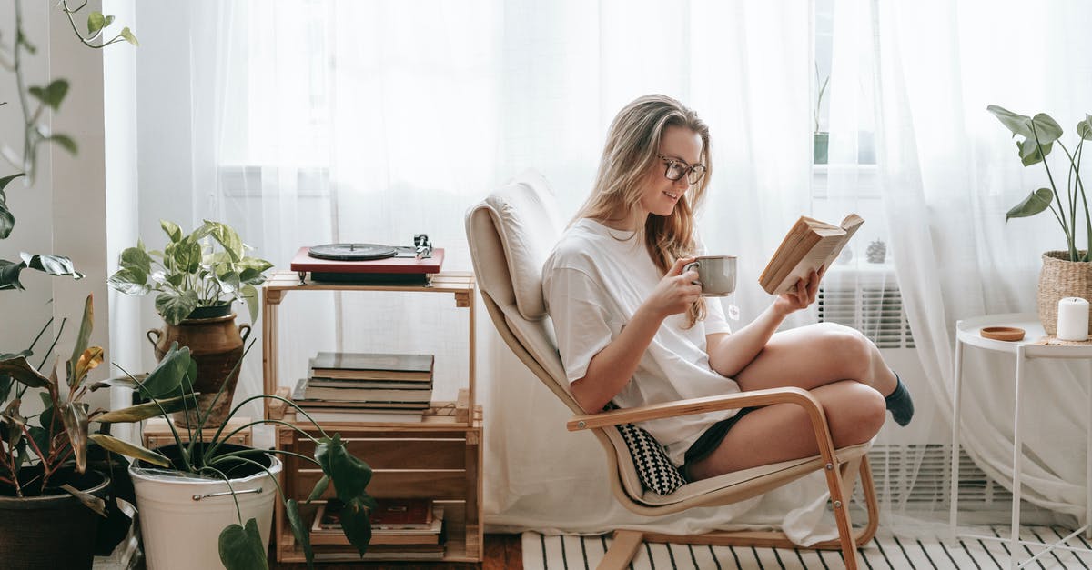 Dragon Age Inquisition Content installing - Cheerful young female in eyeglasses with cup of beverage reading textbook in armchair between potted plants in house room