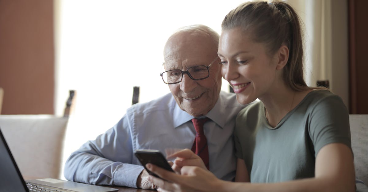 Exp. Share and Pokérus in second generation (GSC) - Smiling young woman in casual clothes showing smartphone to interested senior grandfather in formal shirt and eyeglasses while sitting at table near laptop