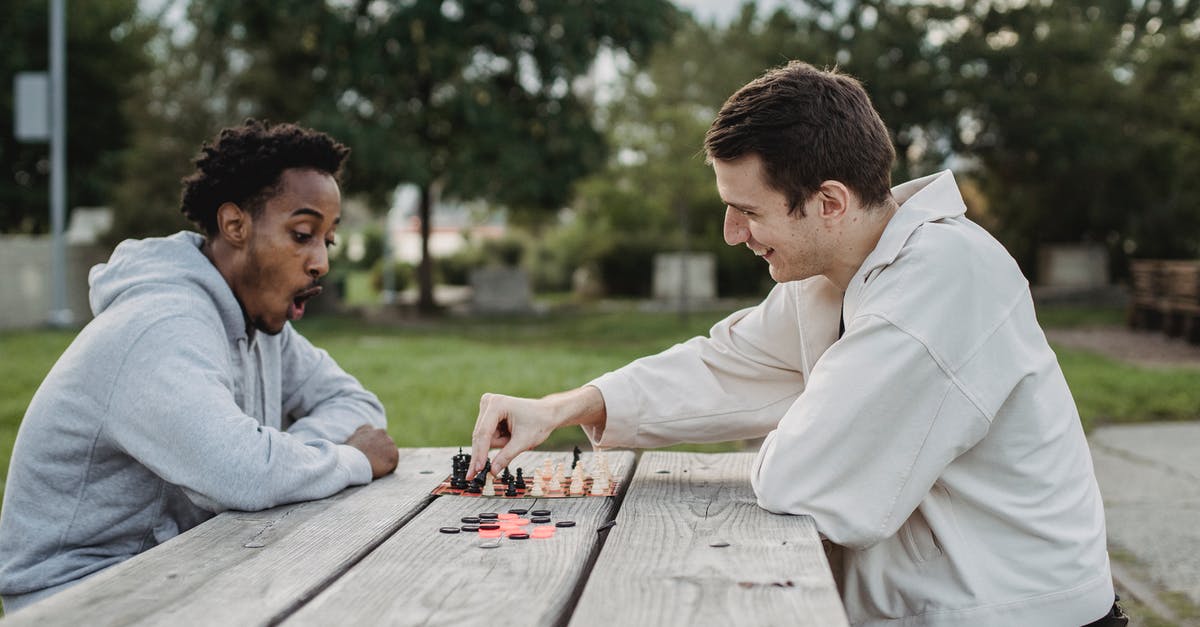 Friendly AI Player Fighting My Allied City State - Side view of smiling young multiracial male friends sitting at wooden table in city park and enjoying interesting chess game