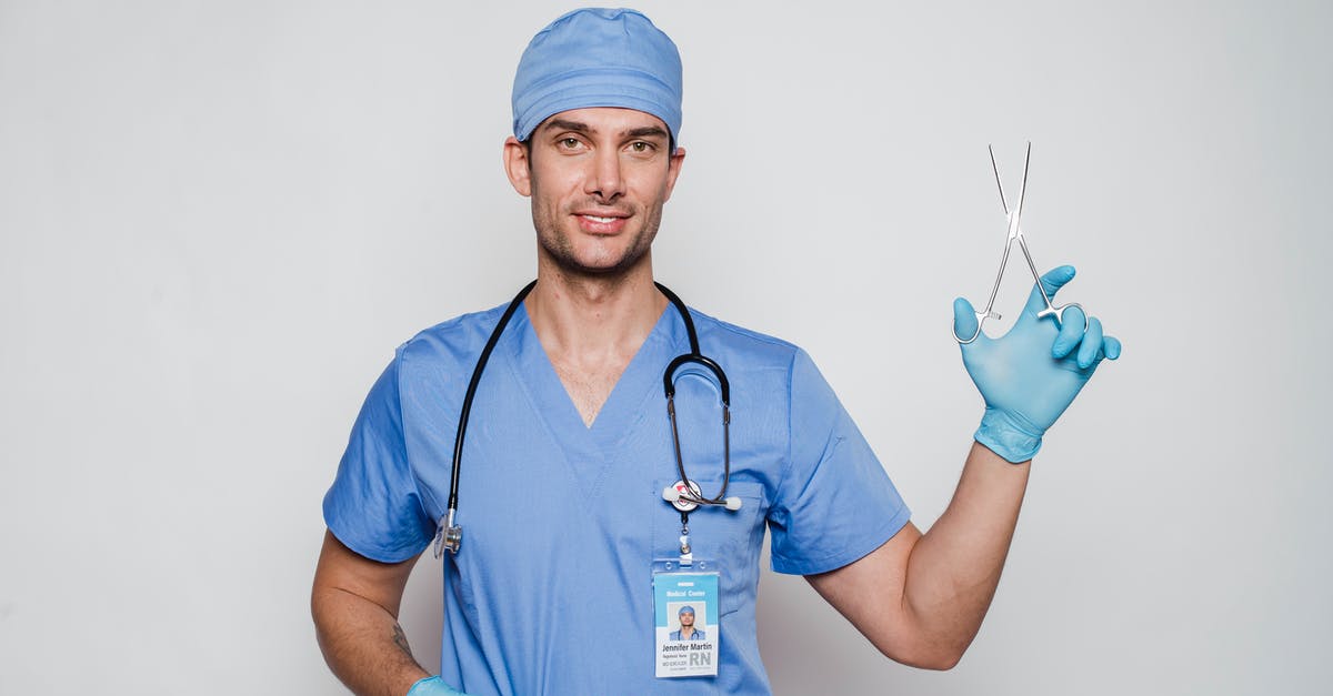 Friendly battle card level cap - Positive adult doctor in blue uniform and cap with gloves and stethoscope with medical instrument and id card looking at camera on white background in light studio