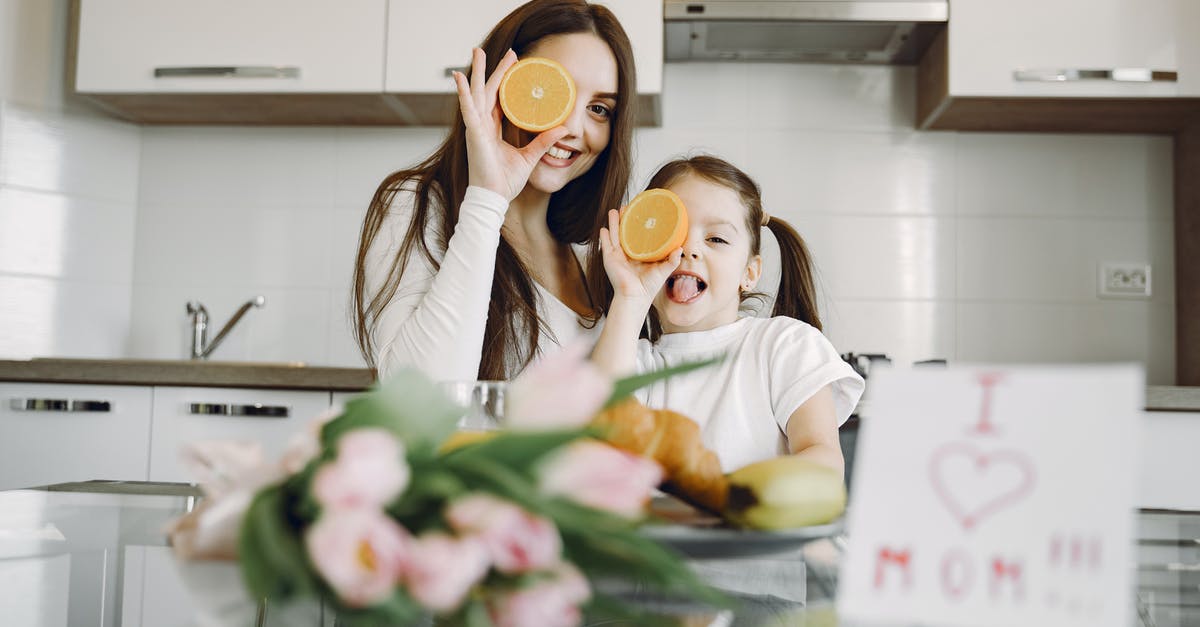 How can I eat whenever I want - From below of cheerful mother and daughter in domestic clothes smiling and playing with oranges while sitting together at table with bouquet of tulip and drawing of child