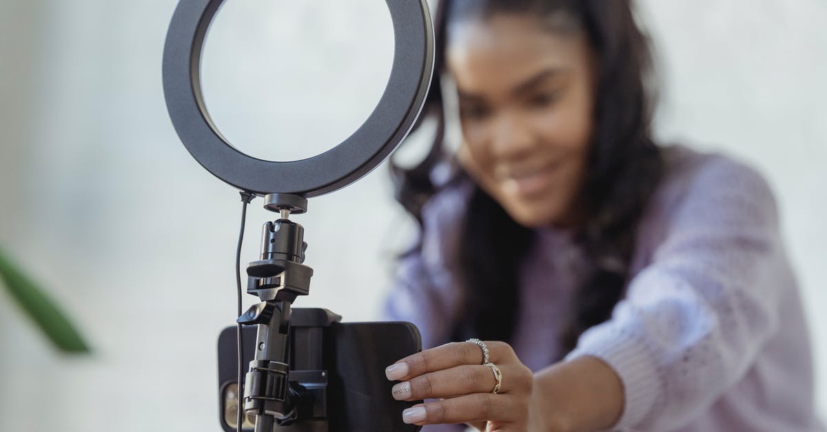 How do I capture a ship? - Cheerful young African American female blogger in stylish sweater smiling while setting up camera of smartphone attached to tripod with ring light before recording vlog