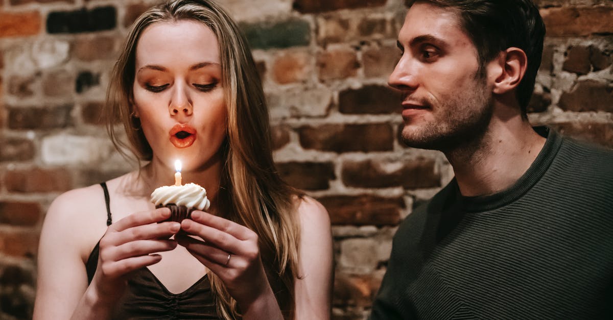 How do I make my friend Notable? - Young couple in elegant outfits in restaurant while blowing in candle on small cupcake and celebrating birthday near brick wall