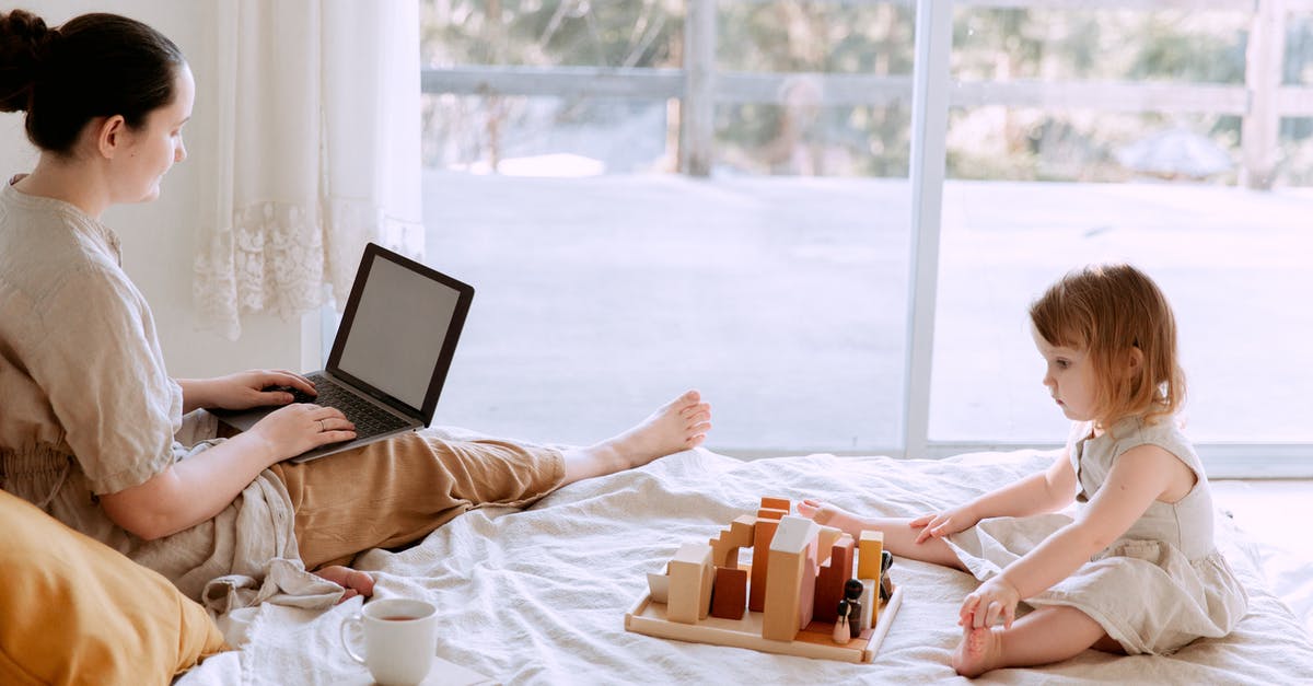 How is a game I haven’t purchased on my home screen? - Side view of casual woman sitting on bed and surfing laptop with blank screen while adorable kid playing with toys in cozy bedroom