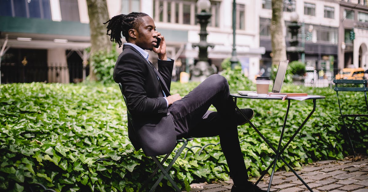 How to chat to all - Side view serious African American businessman in black formal wear sitting at table in city park with legs crossed and having conversation via modern smartphone