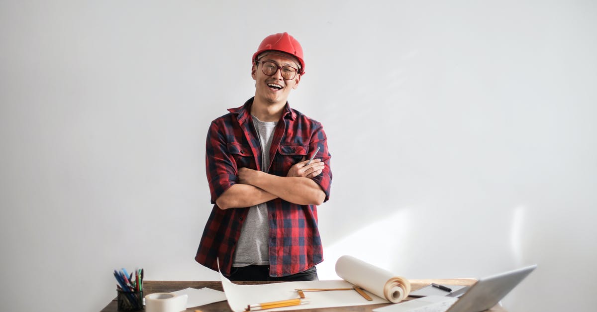 How to develop a buying strategy? - Smiling casual man in hardhat and glasses holding arms crossed looking at camera while standing at desk with paper draft and stationery