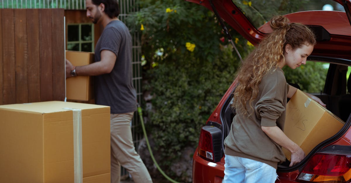 How to get a Red Boundary - Focused young woman with curly hair taking cardboard package out from red automobile trunk on street near stack of moving boxes while bearded ethnic man carrying box into new house