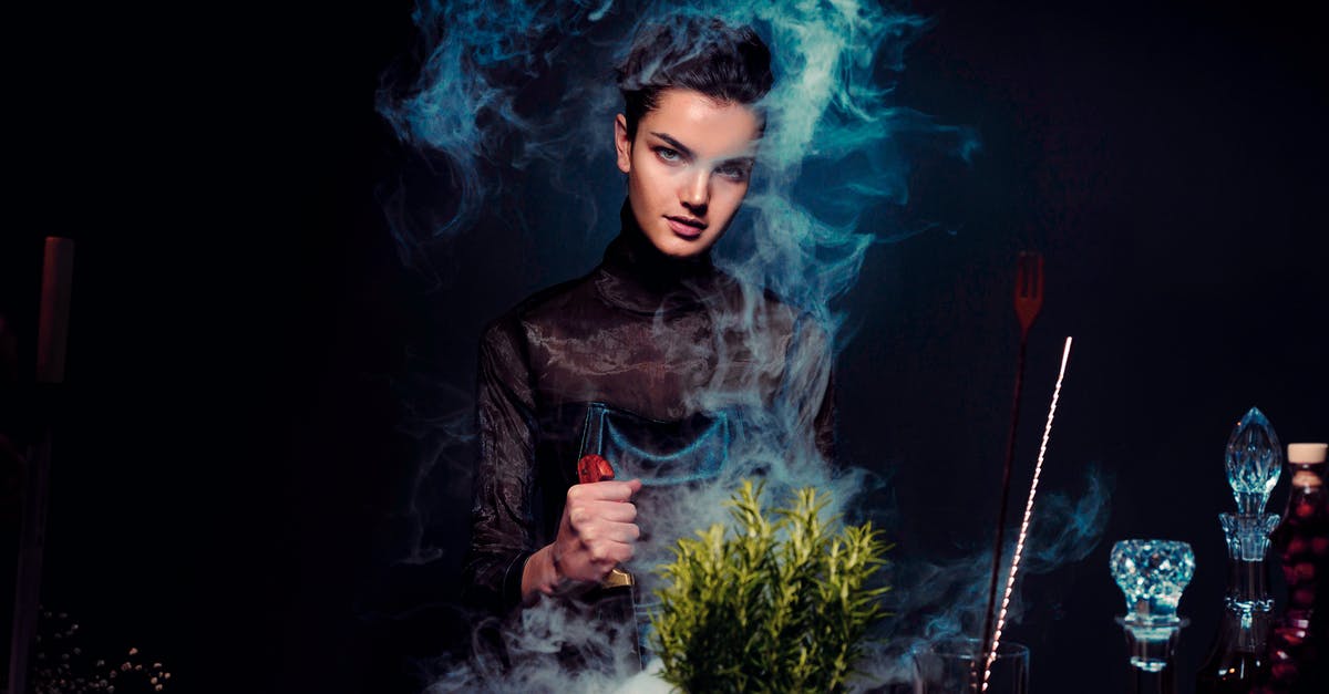 How to make a lingering potion stay longer? - Graceful young female alchemist with knife in hand in black outfit preparing potion from various herbs among smoke in dark room