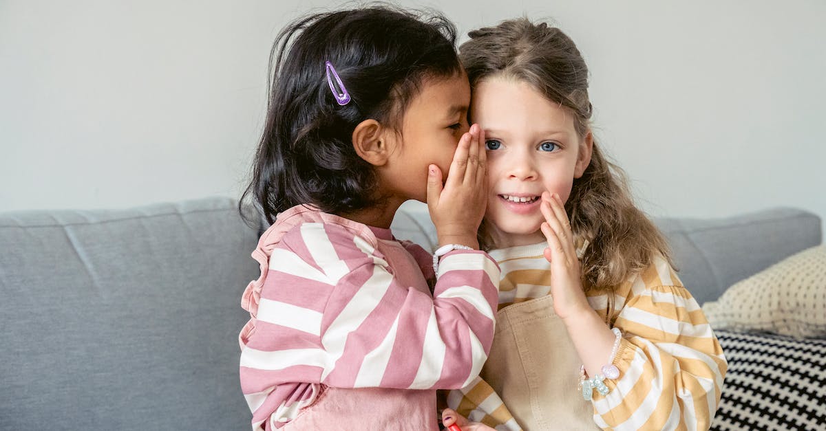 How to tell if I won a challenge? - Ethnic girl sitting on sofa close to smiling best friend and whispering secrets on ear