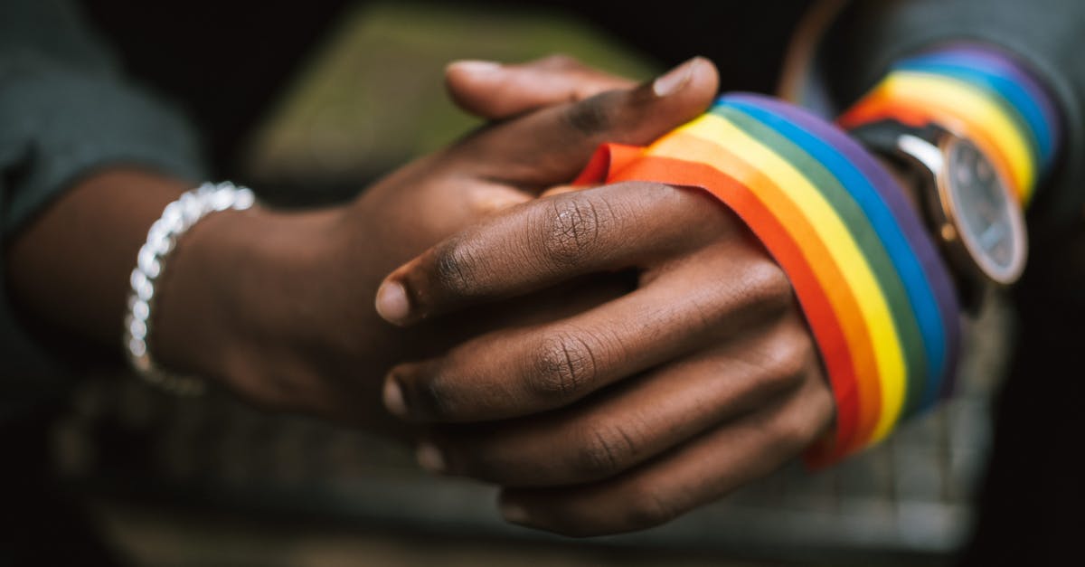 How tolerant are cuccos? - Crop anonymous African American homosexual male wearing colorful rainbow ribbon on arm while sitting on street with hands clasped