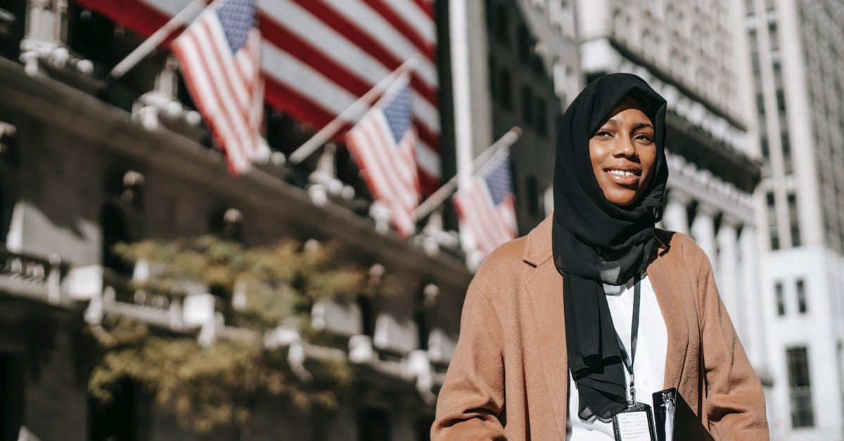 Identifying Uncraftables - From below of cheerful African American female ambassador with folder wearing hijab and id card looking away while standing near building with American flags on blurred background