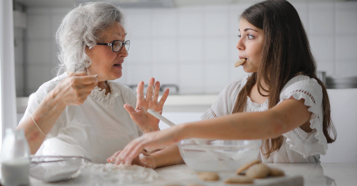 Is it possible to turn off shared games from your friend? - Calm senior woman and teenage girl in casual clothes looking at each other and talking while eating cookies and cooking pastry in contemporary kitchen at home