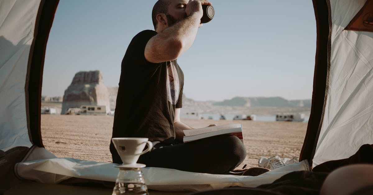 Is my story over? - Side view of male traveler sitting in tent with book near pour over cup while drinking hot beverage in nature during trip