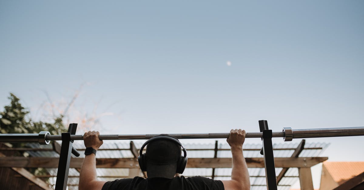 Machines keep using power - Back view of anonymous muscular male athlete listening to music in headphones while exercising on bar under light sky