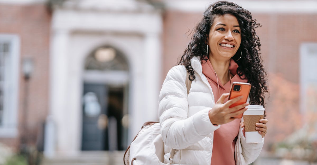 Notifications not going away - Cheerful young ethnic female student with long curly hair in casual clothes and backpack smiling and looking away while using mobile phone standing in campus with takeaway coffee