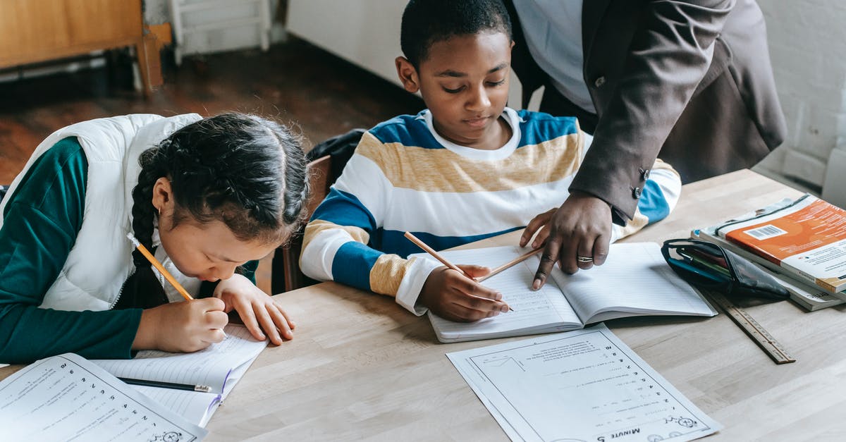 Perk Point maximum - High angle of crop unrecognizable black female teacher explaining task to focused little schoolboy sitting at desk near attentive Asian classmate writing in notebook