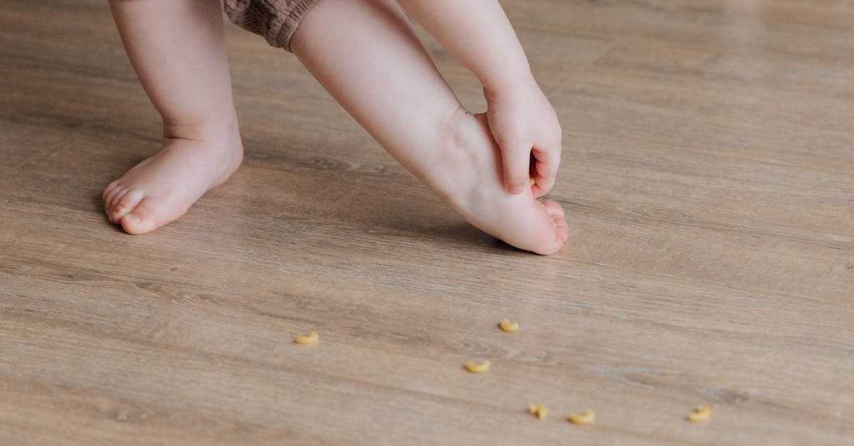 PS3 stuck on 576p - Crop faceless toddler standing barefoot on floor and trying to remove stuck pasta from foot while playing and developing fine motor skills at home