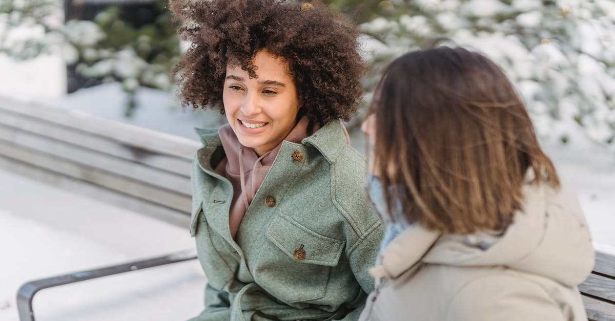 Removing freeze time forever - High angle of positive African American female with curly hair sitting on bench near girlfriend while spending time in winter park