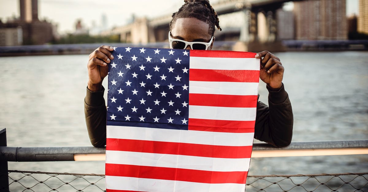 Sirris' sign does not show up on the bridge - Ethnic male in casual clothes and sunglasses standing on embankment of city river while leaning on fence showing national flag of United States of America