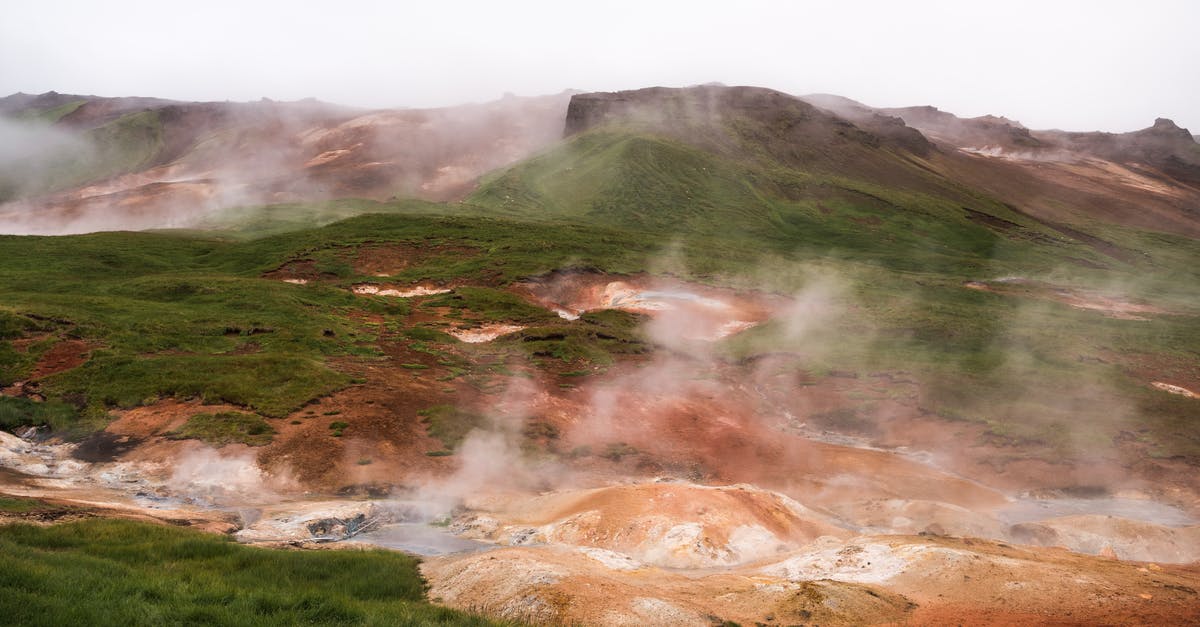 Steam across countries and accounts - Hilled country with geysers blowing steam off among grass area against volcanic mountains in cloudy day