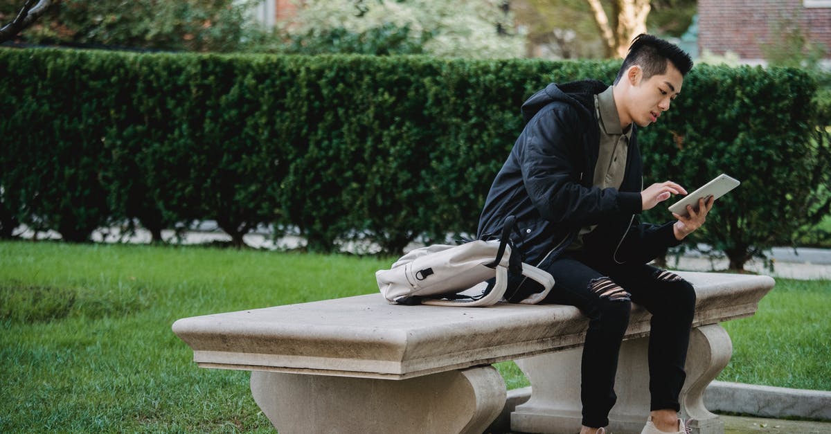 What's the use of fence? - Full body of young focused Asian male in casual wear sitting on bench in park and browsing internet on tablet