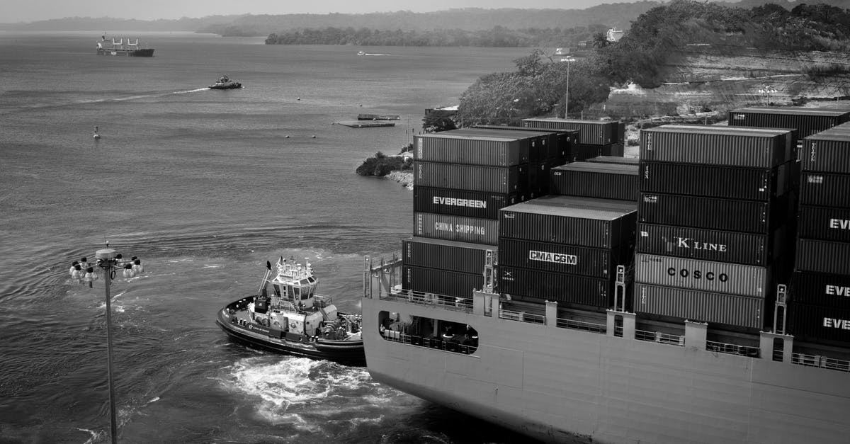 What are the benefits of having a cargo freight station? - Black and white contemporary cargo vessel with many containers floating on calm river near hilly grassy shore towards industrial port