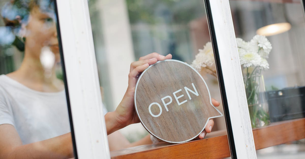 What do I put under the door in the hotel? - Young woman worker in casual clothes standing in light cafe and putting sign with word open on door