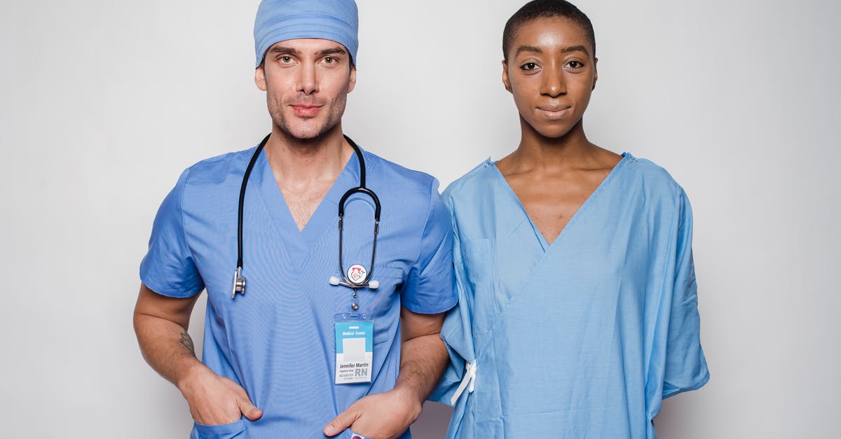 What is this badge? - Positive male doctor in blue uniform with hands in pockets smiling and looking at camera while standing near nurse holding hands behind back in hospital