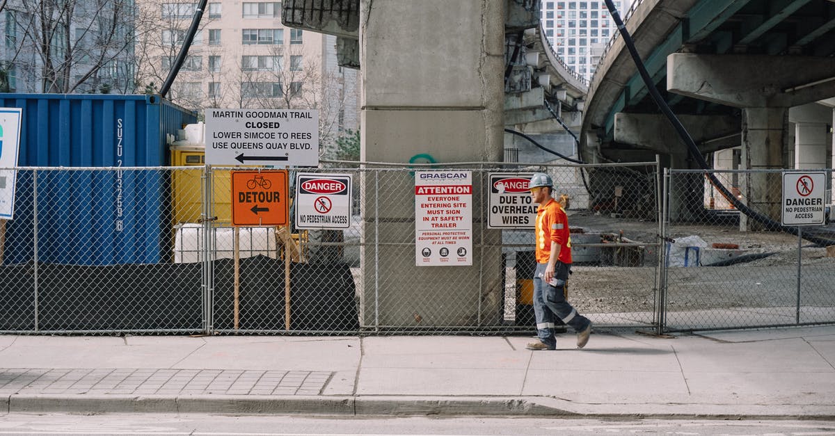 What is this downtown side job during the Corpo prologue? - Full body side view of adult male worker in orange uniform and grey helmet walking under big bridge along chain link fence on street in daylight What is this downtown side job during the Corpo prologue? - Full body side view of adult male worker in orange uniform and grey helmet walking under big bridge along chain link fence on street in daylight