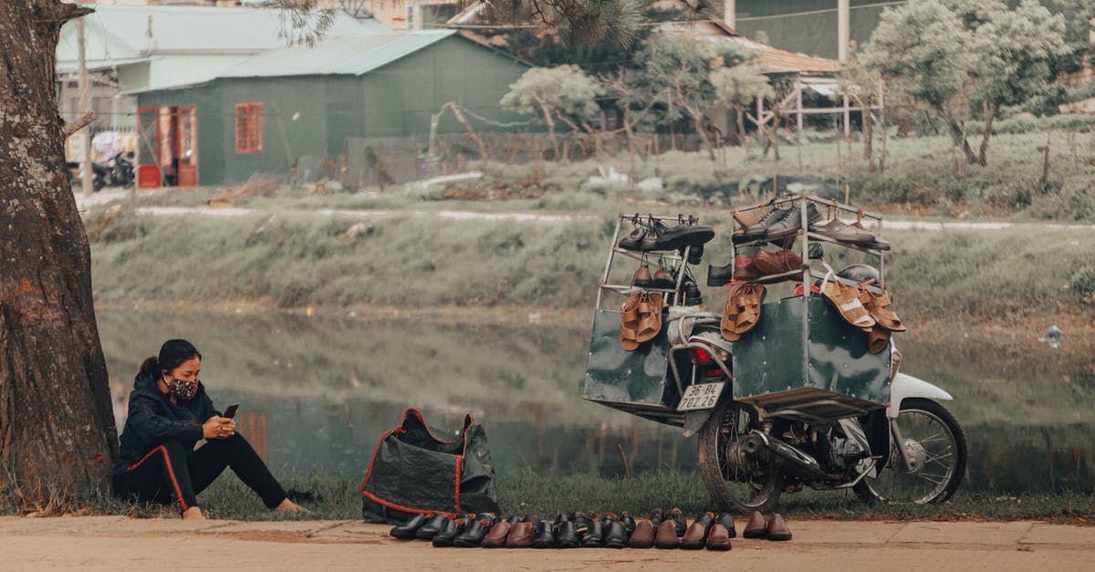 Where are the NPC settlements and vendors? - Full body of focused female street seller in casual clothing and face mask surfing internet on cellphone while selling shoes by road next to pond in settlement