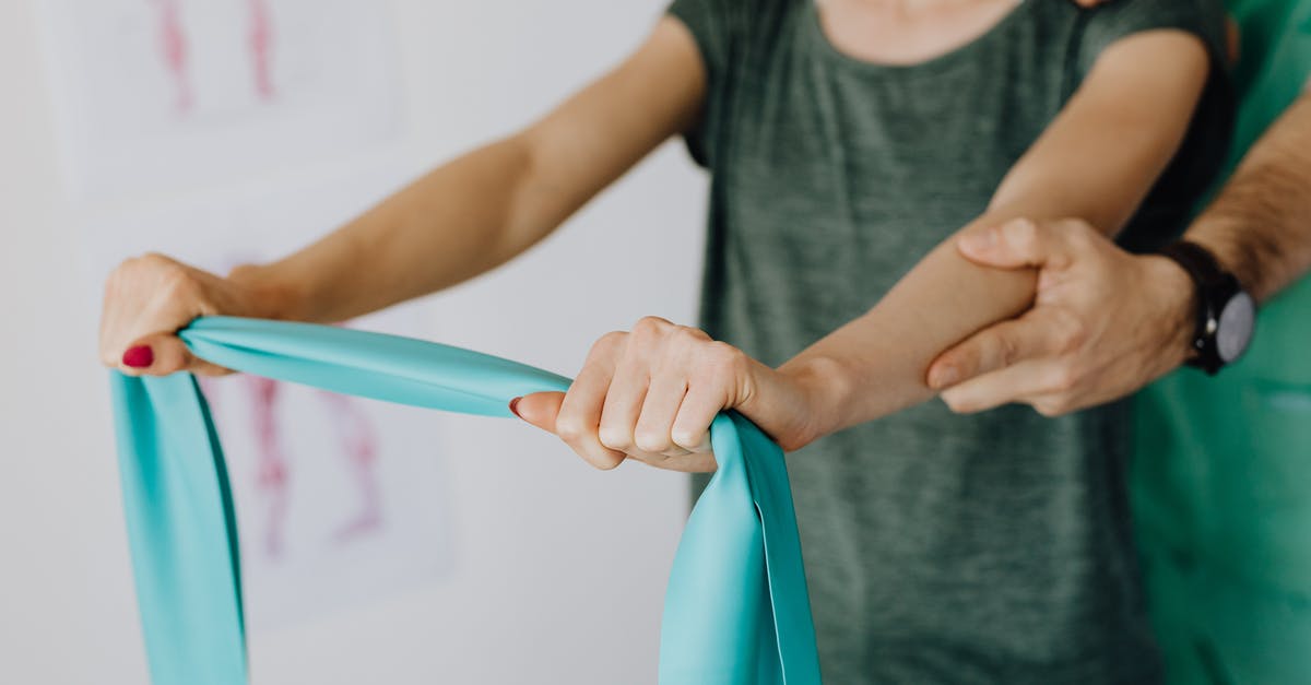 Wilken's Reach and Zunimassas - Crop unrecognizable orthopedist in uniform and wristwatch supporting shoulder and reached arm of faceless female patient with resistance band in doctor office