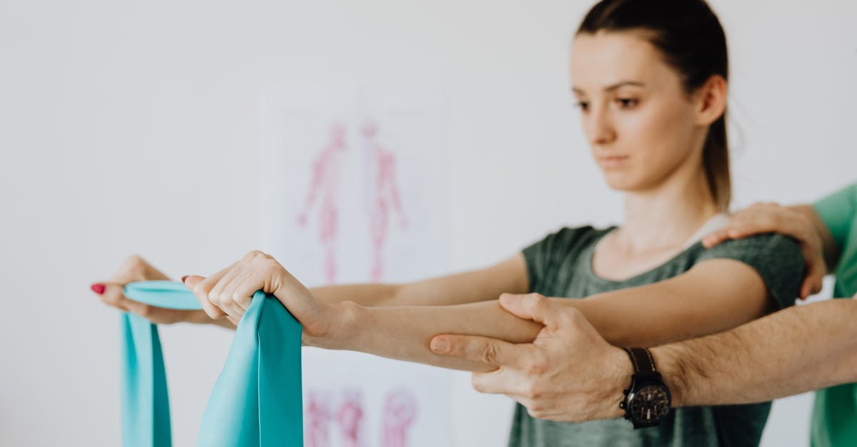 Wilken's Reach and Zunimassas - Slim young female in casual wear standing with reached arms and elastic tape during medical checkup by crop unrecognizable osteopath in wristwatch